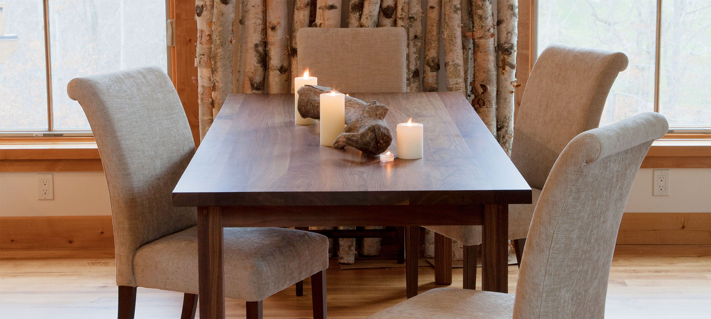 A walnut 4 leg dining table with 4 beige upholstered chairs. There are candles on the table.