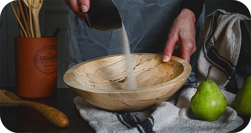 a close up of a woman pouring salt into a large wooden bowl